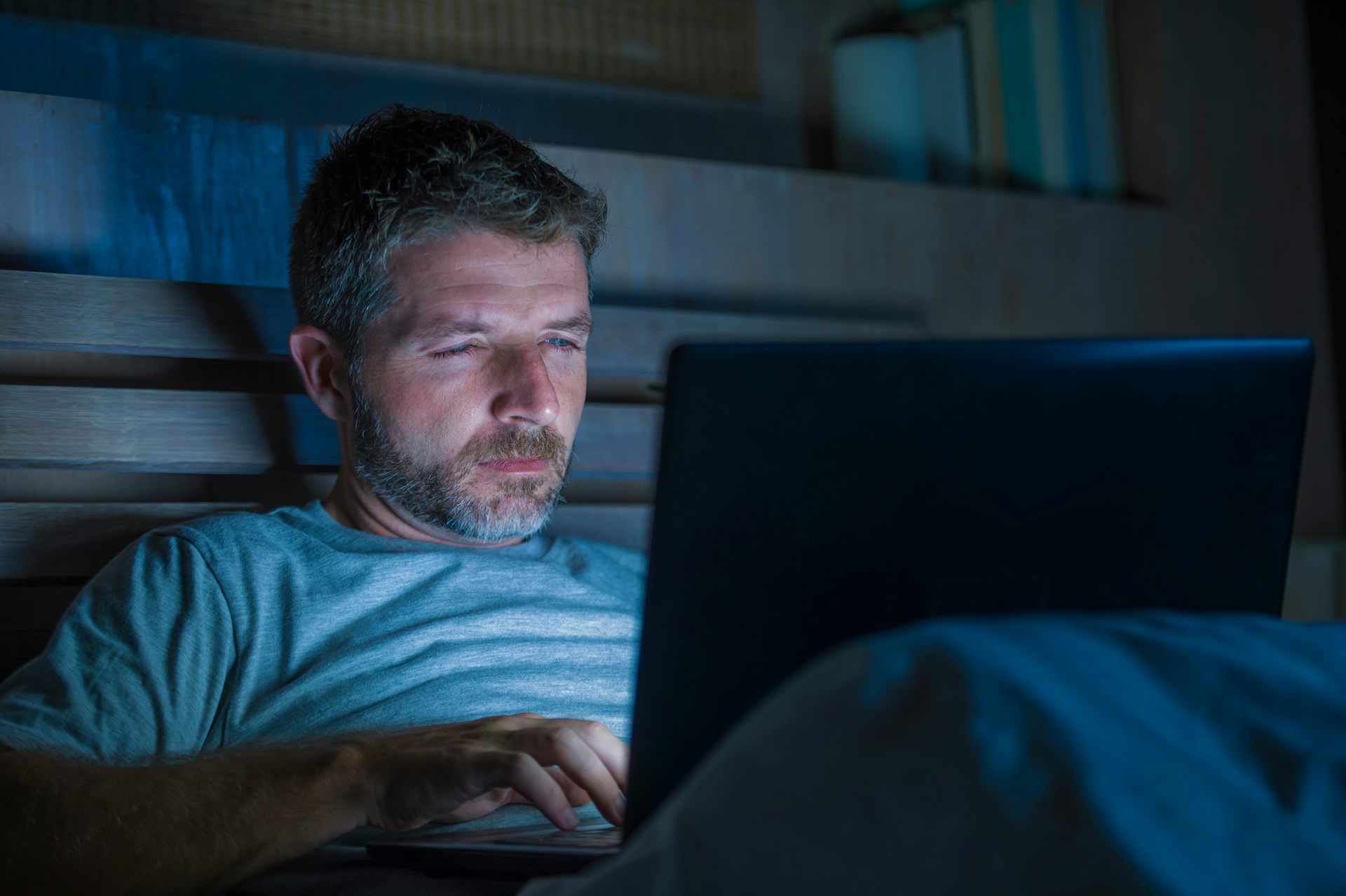 A man wearing a t-shirt sits in bed in a dark room looking at a laptop computer screen